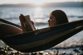 A lovely young girl relaxes in a hammock, reading a book by the lake with golden sunset rays reflecting in the water. 