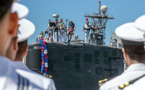 Sailors in white uniforms stand on top of a military submarine as other sailors in white uniforms and hats look on from the foreground.