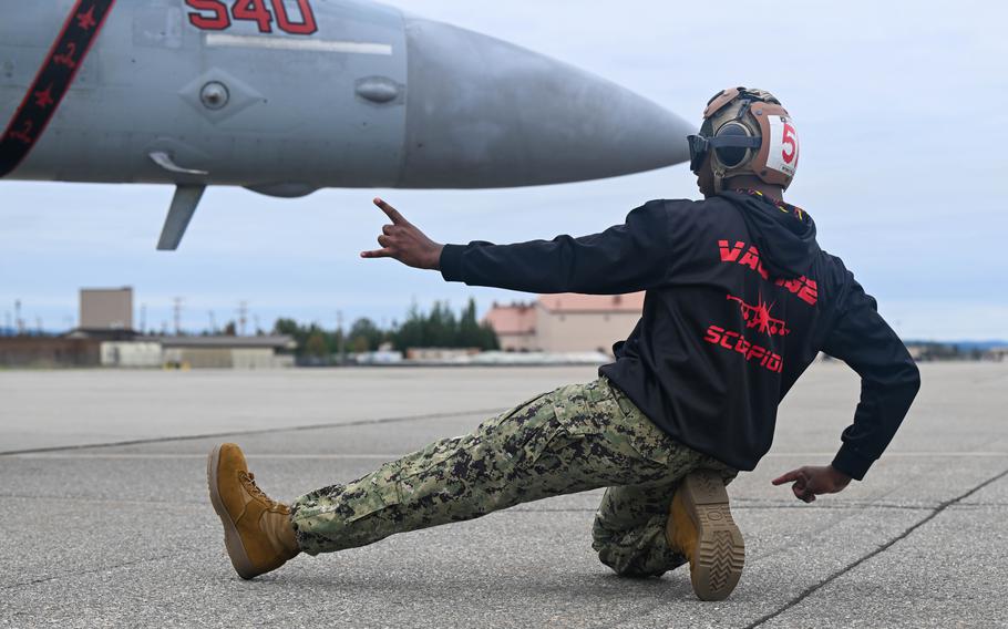 A service member in a sweatshirt crouches and leans back on a runway, gesturing with both hands.