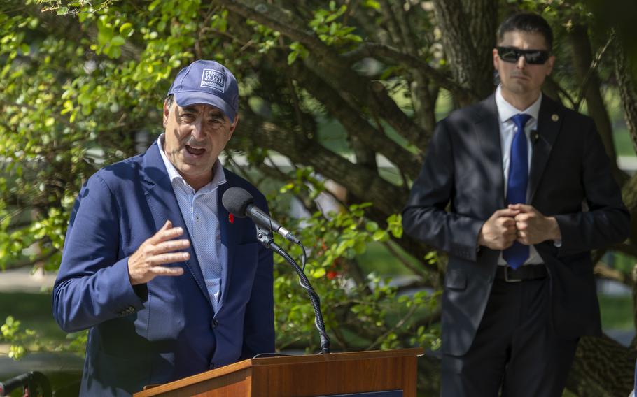 A man in a suit and blue hat speaks at a wooden podium, gesturing with one hand.