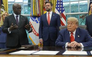 A Rwandan government official speaks in the Oval Office of the White House while standing next to a desk where Donald Trump is seated listening. JD Vance stands between them listening.