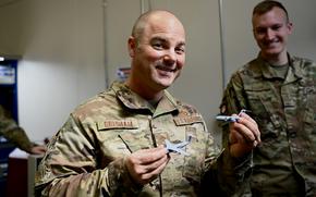 A man in a camouflage military uniform smiles while looking forward and holding up a small aircraft in each hand.