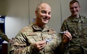 A man in a camouflage military uniform smiles while looking forward and holding up a small aircraft in each hand.
