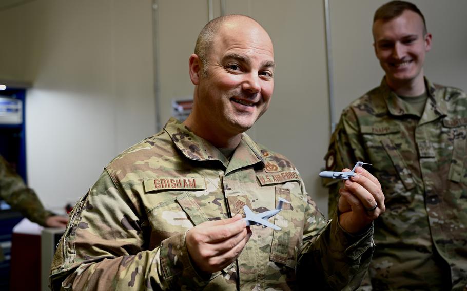 A man in a camouflage military uniform smiles while looking forward and holding up a small aircraft in each hand.