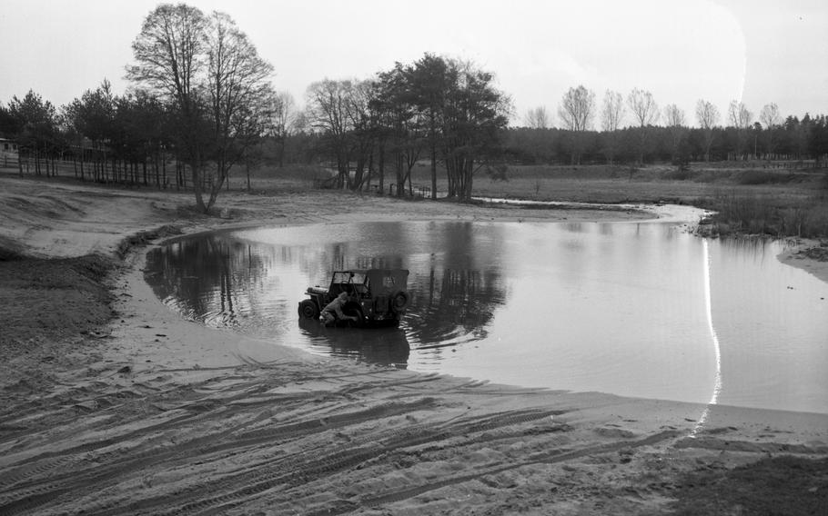 A black and white photo of a military Jeep in a pond, with a soldier crouching to check on it.