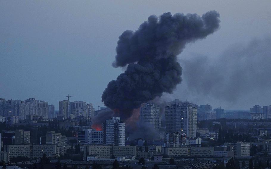 A large plume of dark smoke, seen from a distance, rises over a city skyline in the early morning light.
