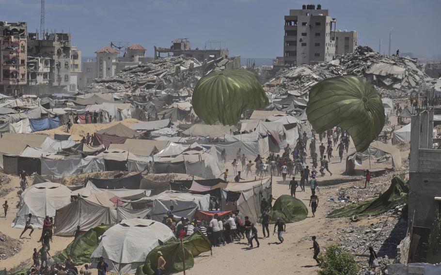 An aerial view of crates attached to parachutes descending into an encampment of tents, with a skyline of damaged and destroyed buildings in the background.