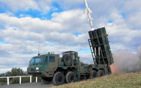 A military missile defense system mounted on a green camouflaged heavy-duty truck is positioned on grass beside a road with a white fence. The system features a large vertical launcher assembly with multiple missile tubes, and a gray missile can be seen launching upward with a visible exhaust plume against a cloudy blue sky. 