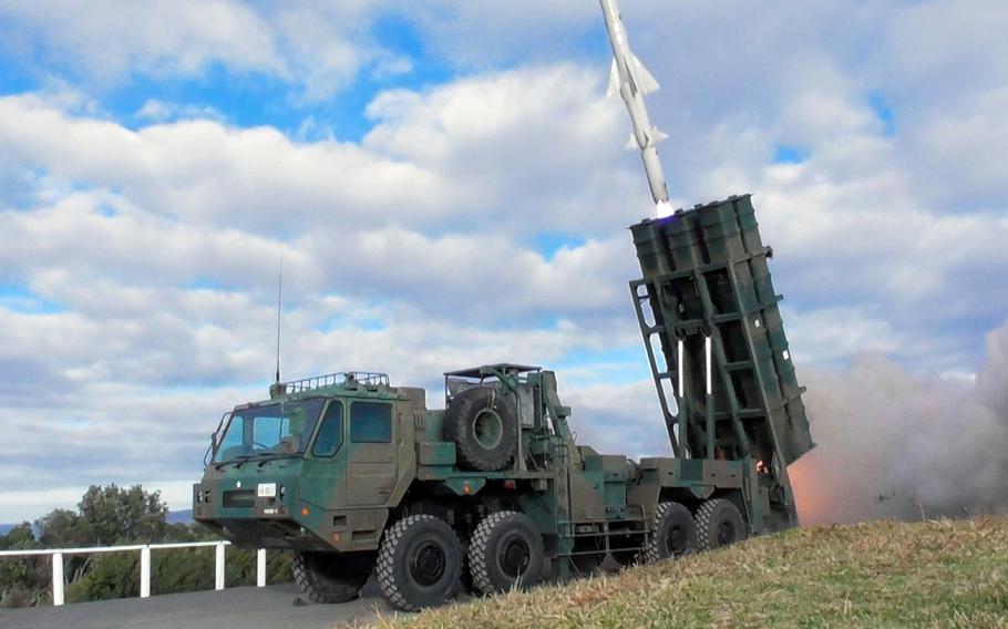 A military missile defense system mounted on a green camouflaged heavy-duty truck is positioned on grass beside a road with a white fence. The system features a large vertical launcher assembly with multiple missile tubes, and a gray missile can be seen launching upward with a visible exhaust plume against a cloudy blue sky. 