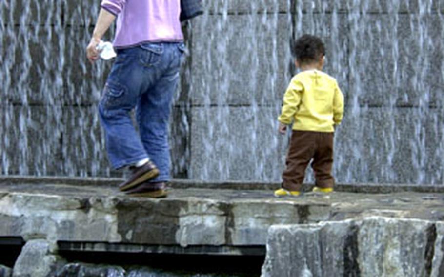 A woman and toddler make their way across a narrow bridge, with a waterfall on one side and the Cheonggye Stream on the other.