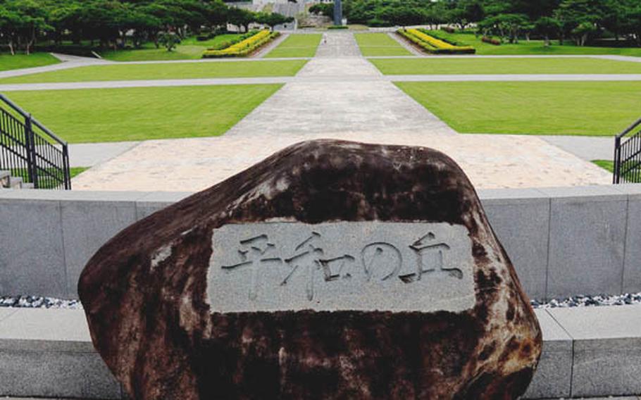 A boulder inscribed with “Hill of Peace” sits at the base of a monument. In the distance is the Peace Memorial Hall where a large Buddha statue is located.