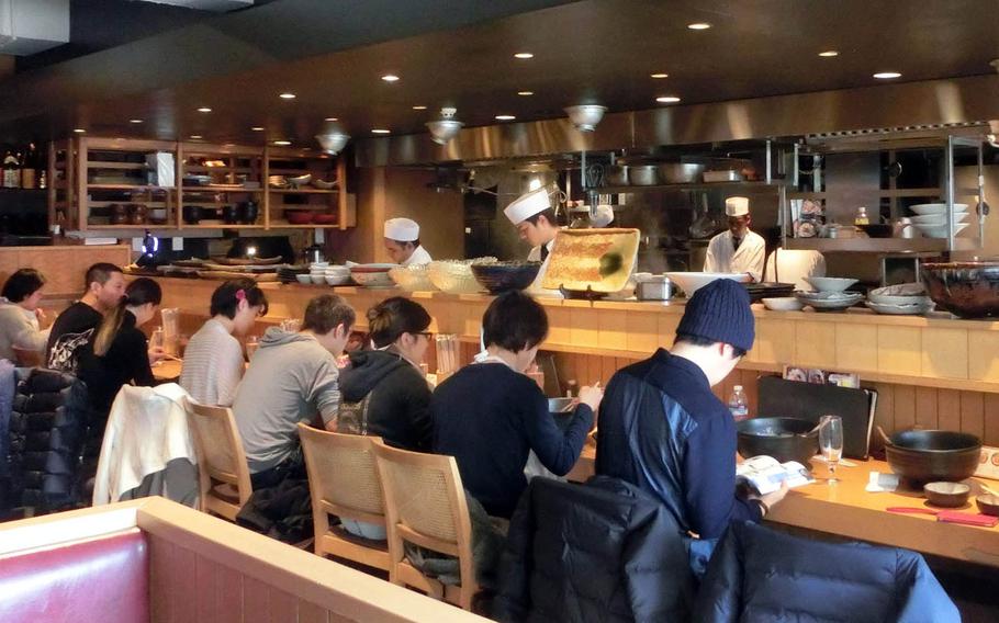 Chefs at Tsuru Ton Tan in Tokyo prepare udon in an open kitchen while customers fill the counter seats.