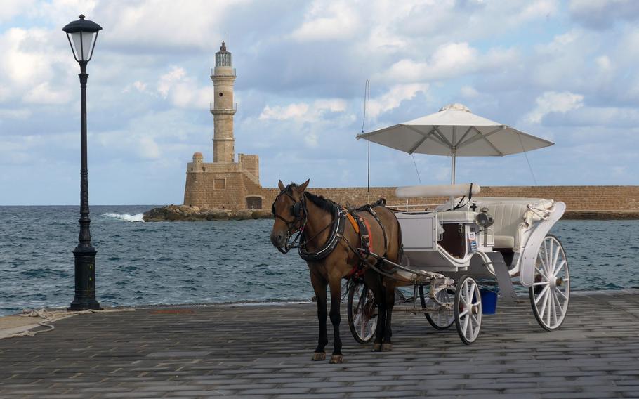 A horse and buggy stands on the Chania Crete's Old Harbor promenade. In the background is the Venetian Lighthouse