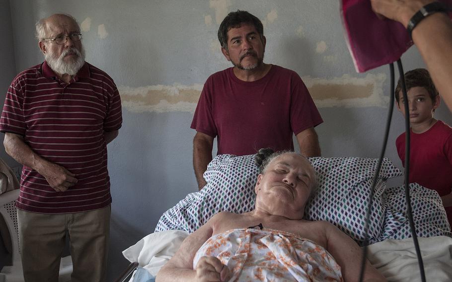 Vietnam veteran Javier Morales, left, looks on with family members of a bed-ridden elderly woman who is about to have a medical check-up by a Red Cross worker in the Ciales region of Puerto Rico, on Nov. 12, 2017.