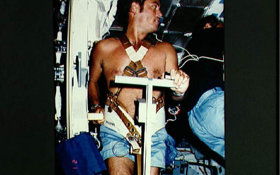 On middeck in front of open airlock hatch in 1983, commander Robert Crippen, restrained by harness, exercises on treadmill as fellow crew members conduct other activities around him.