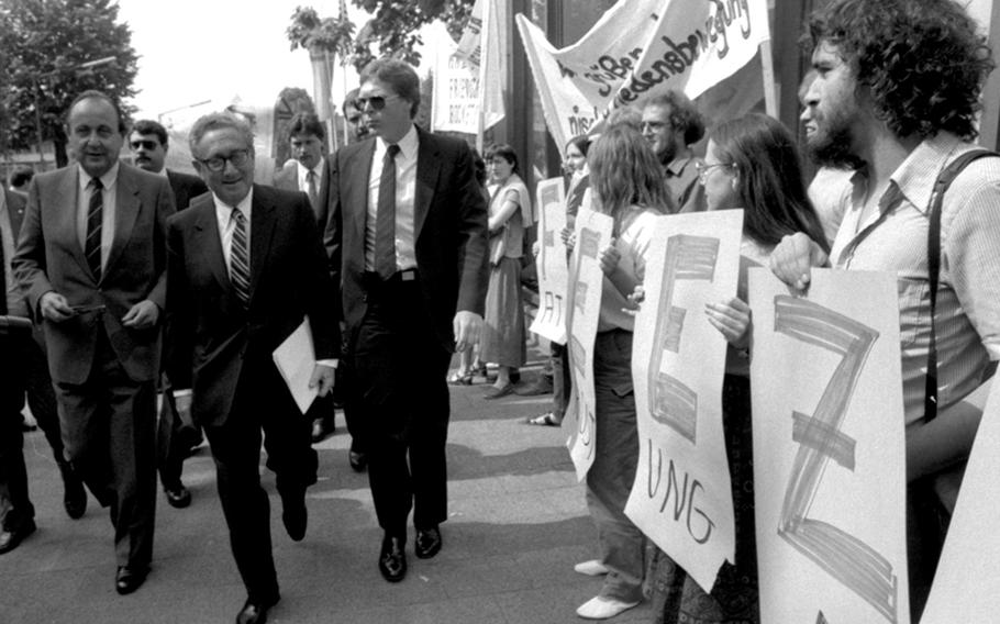 Former Secretary of State Henry Kissinger walks past a line of protesters on his way to speak at a German-American tricentennial ceremony at Worms, Germany, in 1983. At left is German Foreign Minister Hans-Dietrich Genscher. The protesters are holding letters forming the word "freeze," in connection with nuclear disarmament.