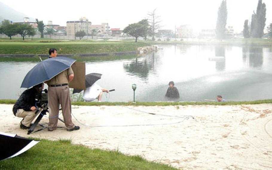 Waist-deep in the island hole at Camp Walker Golf Course in Taegu, South Korea, Army Col. James M. "Mike" Joyner films a TV spot with safety tips for Korea's monsoon season. Visible at right, in pond, is Kevin B. Jackson, Joyner's public affairs chief.