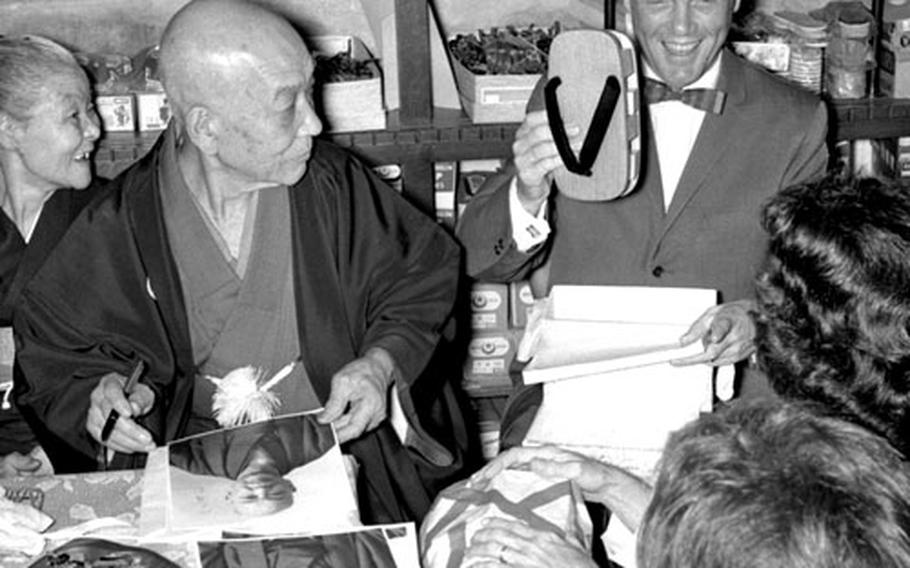 Tokuzo Shibuya and his wife watch as astronaut John Glenn unwraps their gift, a pair of wooden clogs.