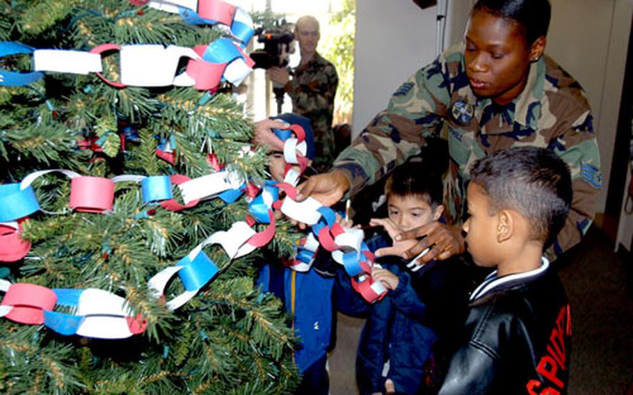 Staff Sgt. Tessiah Hemphill, Family Readiness noncommissioned officer at the Family Support Center, helps Apollo Morales, foreground, and Javier Guzman drape a paper train on a Christmas tree in the Torii Building lobby Thursday morning. About 60 students from Sollars Elementary School with a parent deployed helped decorate the tree with photographs of their mom or dad and other handmade ornaments.