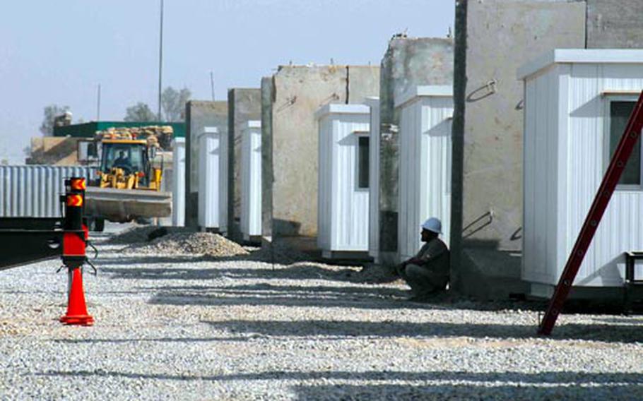 A worker at Forward Operating Base Delta rests in the shade of a row of new Contained Housing Units, or one-room trailers, as another worker moves one of the concrete barrier walls that will protect this "CHU city" from explosives.