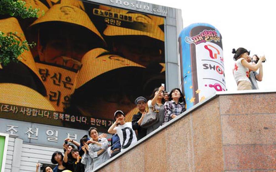 Mourners watch a procession carrying the body of former South Korean president Roh Moo-hyun through Seoul on Friday. Behind them is a screen showing other mourners outside Seoul City Hall wearing yellow visors. South Korean politicians or parties are typically represented by a color, and yellow came to be associated with Roh during his political career.