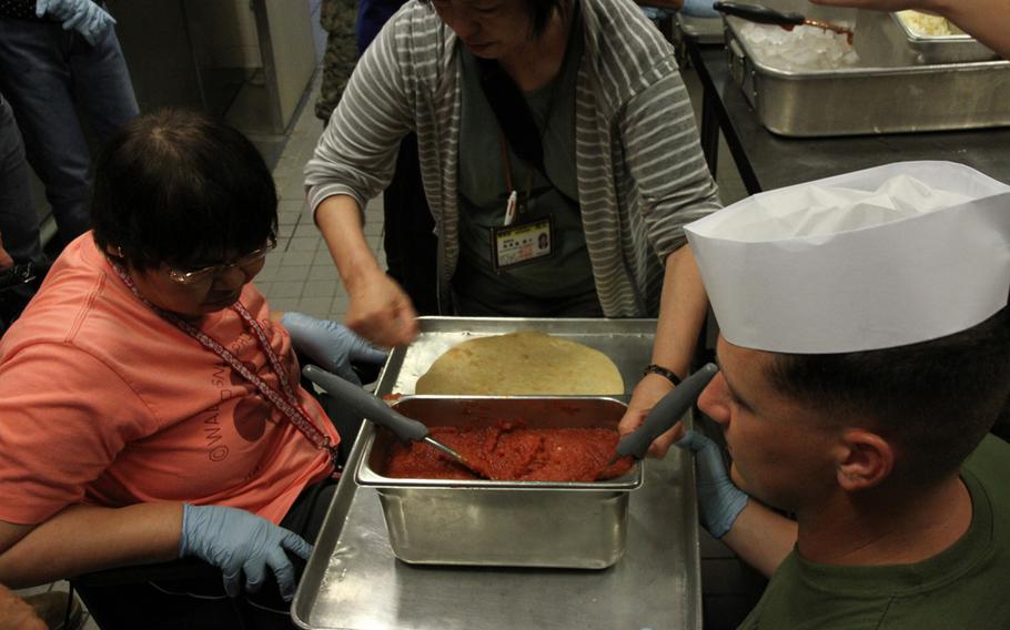Marine Sgt. Ryan Tipton takes kneels to help show a resident from the Reimei No Sato home for disabled adults how to make a pizza on Thursday, July 7, 2016 at the 12th Marines Mess Hall on Camp Hansen. The residents learned to make their own pie and then got to sit with the Marines and taste what they had made. They also made arts and crafts.