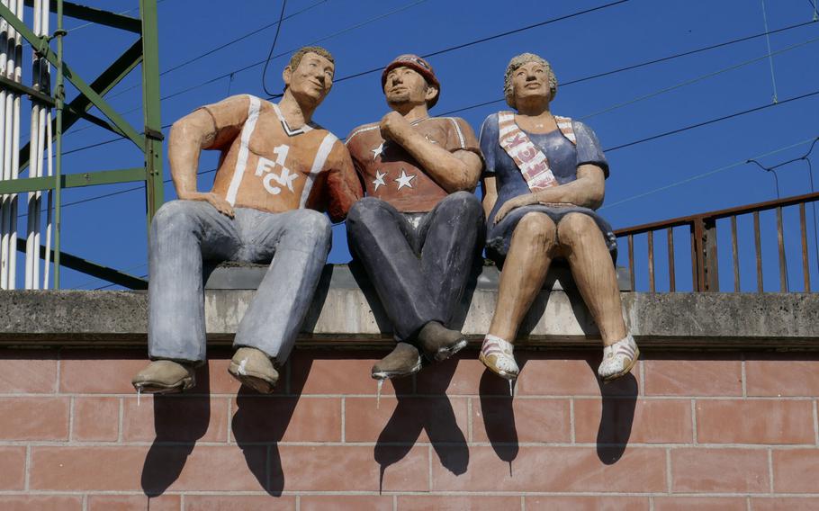 Three 1. FC Kaiserslautern fans sit on a wall near the train station, looking down at a team that is part of an installation on a traffic circle at the foot of the road leading to the legendary Fritz Walter Station, home to the city's popular soccer team.