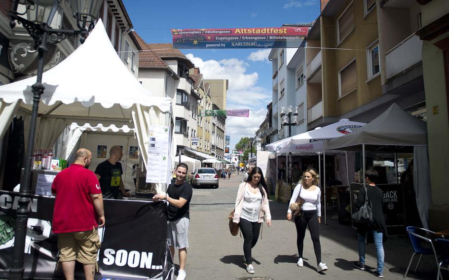 Two people secure a sign to a white booth as pedestrians walk by. Overhead is a large red and black banner advertising the Altstadtfest. Other booths can be seen in the background. 