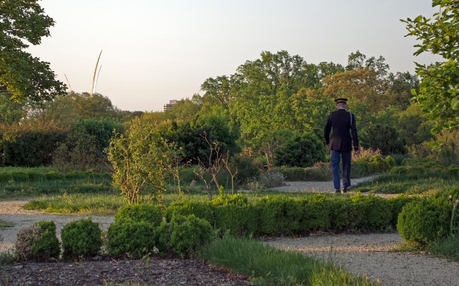 A servicemember walks through the flower garden at the Arlington House shortly after the sunrises on May 4, 2015, in Arlington National Cemetery. Through the trees is the top of the Air Force Memorial. 