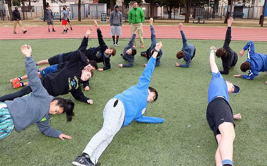 Yokota track and field athlletes warm up under the watchful eye of coach Dan Galvin, background.