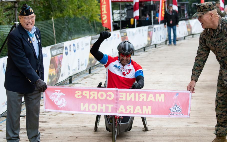 Omar Duran crosses the finish line at the 43rd Annual Marine Corps Marathon, held throughout Washington, D.C., on Sunday, Oct. 28, 2018. Holding the banner is Medal of Honor recipient Hershel "Woody" Williams and Marine Corps Commandant Gen. Robert Neller.