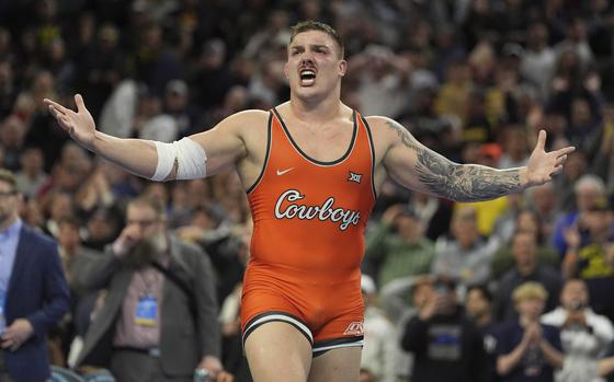 A wrestler in an orange singlet reading “Cowboys” spreads his arms in celebration in front of a large crowd at a meet.