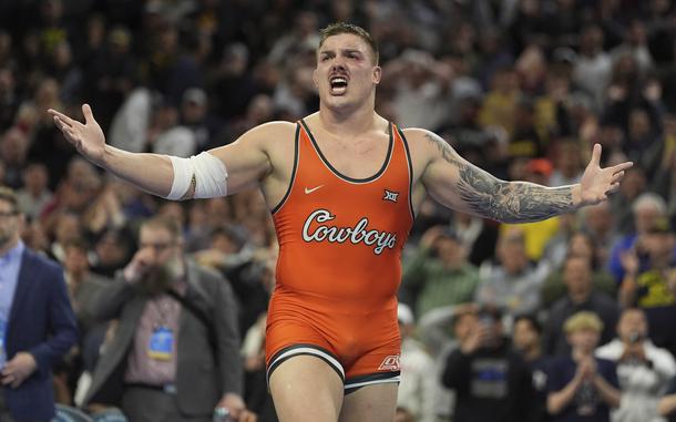 A wrestler in an orange singlet reading “Cowboys” spreads his arms in celebration in front of a large crowd at a meet.