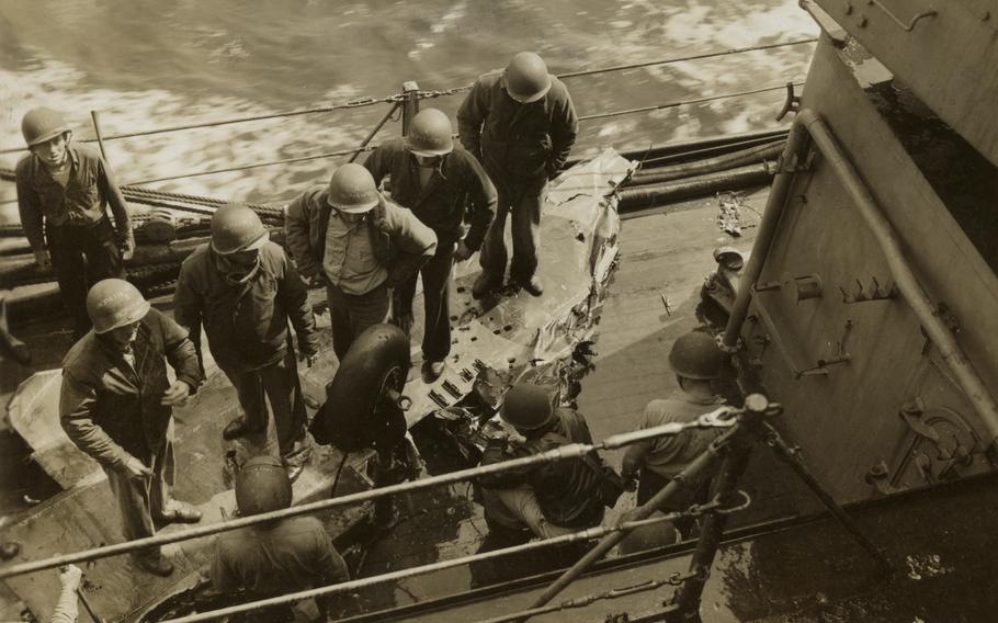 A black-and-white, 1940s-era image of military members, seen from above, standing near the rail of a ship at sea and surveying wreckage of an airplane.