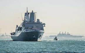 The USS New Orleans, a San Antonio-class amphibious transport dock, navigates through waters with a smaller patrol boat in the foreground and additional naval vessels visible in the background. 