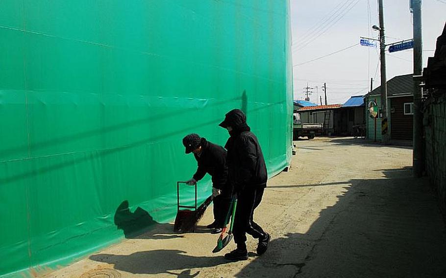 Two women sweep the road outside a green tarp that houses the shells of several buildings destroyed by a North Korean attack in 2010. The women are part of a community improvement group paid by the government to clean the village streets.