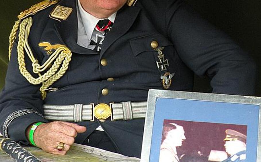 A gentleman portraying a World War II-era German officer sits behind a photo of the officer he portrays. The officer in the photo is shaking hands with Adolf Hitler. 