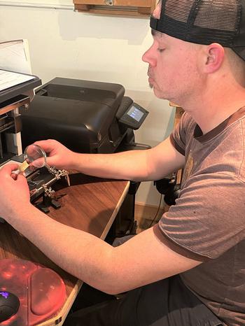 Former Army Capt. Andy Reed looks through a magnifying glass as he engraves a plaque to go on a presentation box for a first salute coin, April 28, 2021. 
