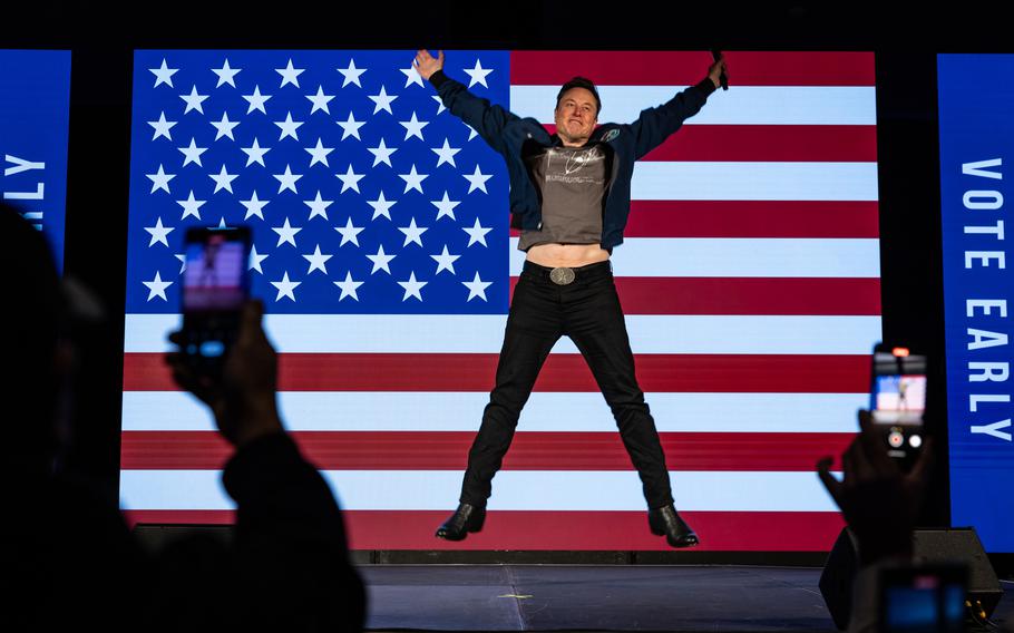 Elon Musk jumps in front of an American flag background while on stage.