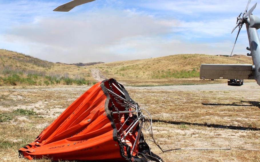 The  bucket carried by the MH-60S Knight Hawk holds about 420 gallons of water, weighing more than 3,000 pounds. The bucket is attached to the helicopter with a cargo hook and allows the aircraft to pick up water from a river or the ocean and dump it on a wildfire. Smoke visible behind the helicopter is from one of three wildfires currently burning at Camp Pendleton, Calif.