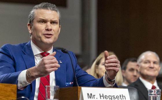 Pete Hegseth gestures with both arms extended while speaking into a microphone.