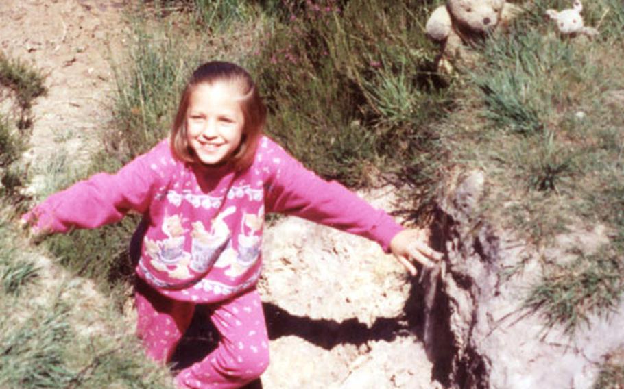 A young girl is all smiles as she climbs up from the dirt and pond that make up the Sandy Pit Where Roo Plays. To her left are the two stuffed Pooh bears.