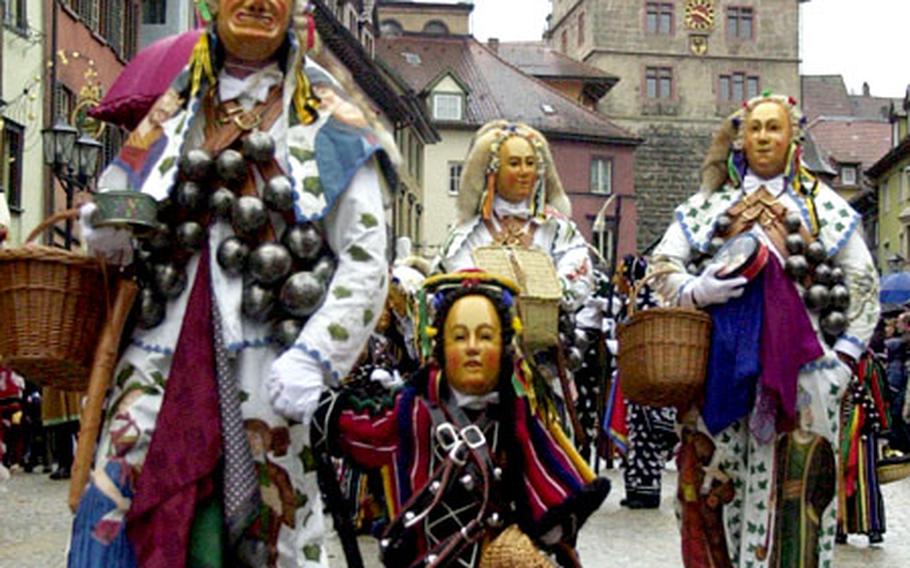 The “Narrensprung” (Fools Jump) begins in Rottweil, Germany. Characters wear a variety of costumes. At right, a tall “Biss” walks with a young “Fransenkleid,” followed by two “Gschell.” In the background is the Schwarzen Tor (Black Gate), where the parade begins.