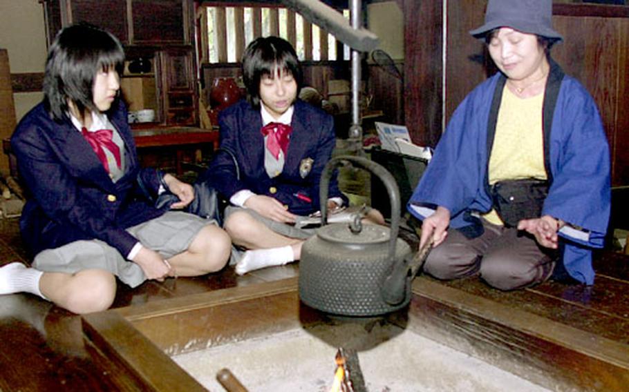 Two girls talk with a volunteer at the Edo-Tokyo Open Air Museum about the old farm house around an irori, an open hearth. The smoke helps to keep insects from destroying the dense hay on the roof.