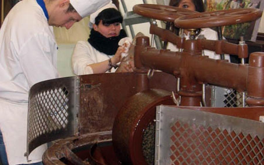 While the machine performs "conching," a crucial step in the making of chocolate that removes excess moisture and eliminates bitterness or acidity, workers in the background prepare molds for making chocolate designs.