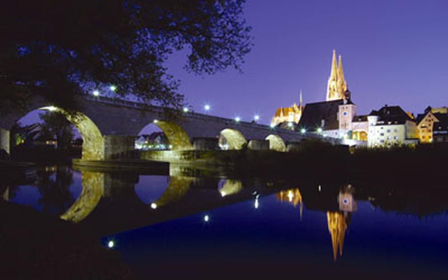 The illuminated Regensburg cathedral is reflected in what is famously known as the “Blue Danube” at twilight. In the foreground is the historic Steinerne Brücke, or Stone Bridge.