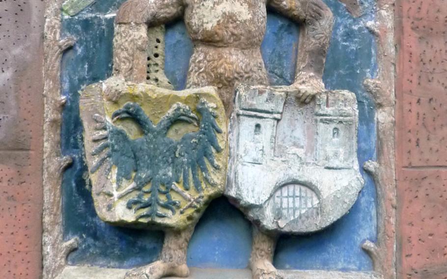 A figure holding a pair of coats of arms hangs on a building in Wissembourg, France. The town's German name, Weissenburg, or white castle, is depicted in the coat of arms on the right, the one on the left is that of the German Confederation.