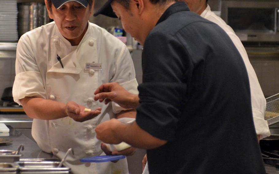Kitchen staffers go about their business at Roy's Hawaii in Okinawa. All except the kitchen staff are dressed immaculately in white tops, black trousers and ties, and the tables are adorned with white tablecloths, blue napkins and gleaming silverware and crystal.