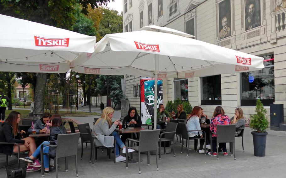 People enjoy a drink at a cafe/bar on Piotrkowska Street, under the watchful eyes of famous people of Lodz, Poland, looking out of widows painted on the facade of the building. Because it is next to a park, the house is known as the Tenement House Under the Chestnuts.