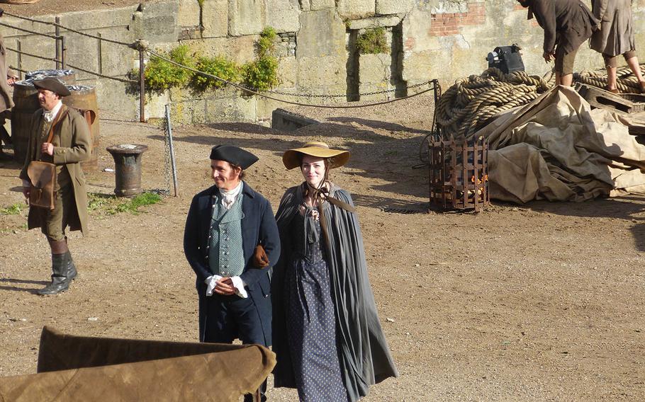 A young woman and a gentleman in a tricorn hat saunter along the harbor in Charlestown.